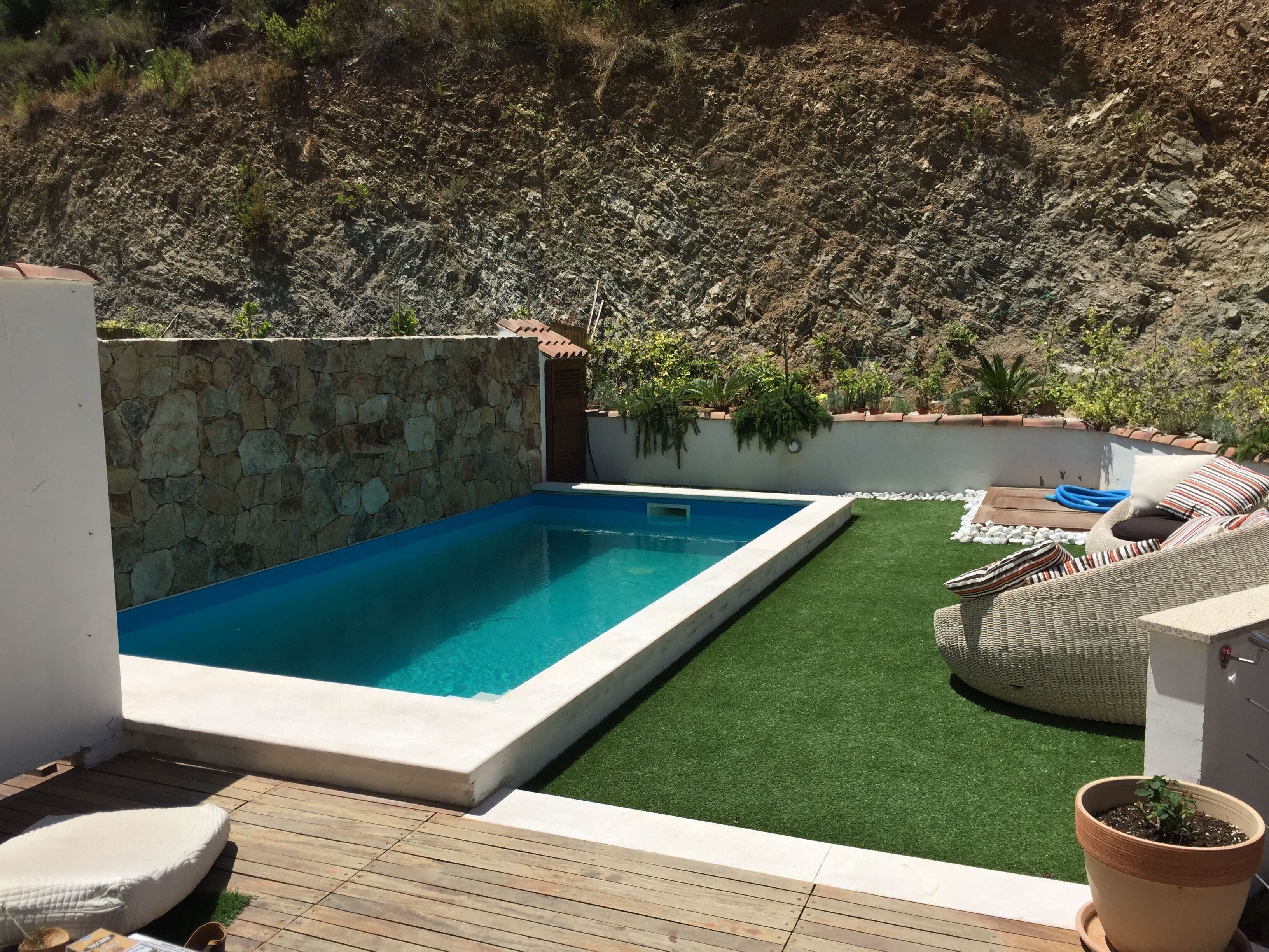 A swimming pool with a stone wall, artificial grass, and outdoor furniture.