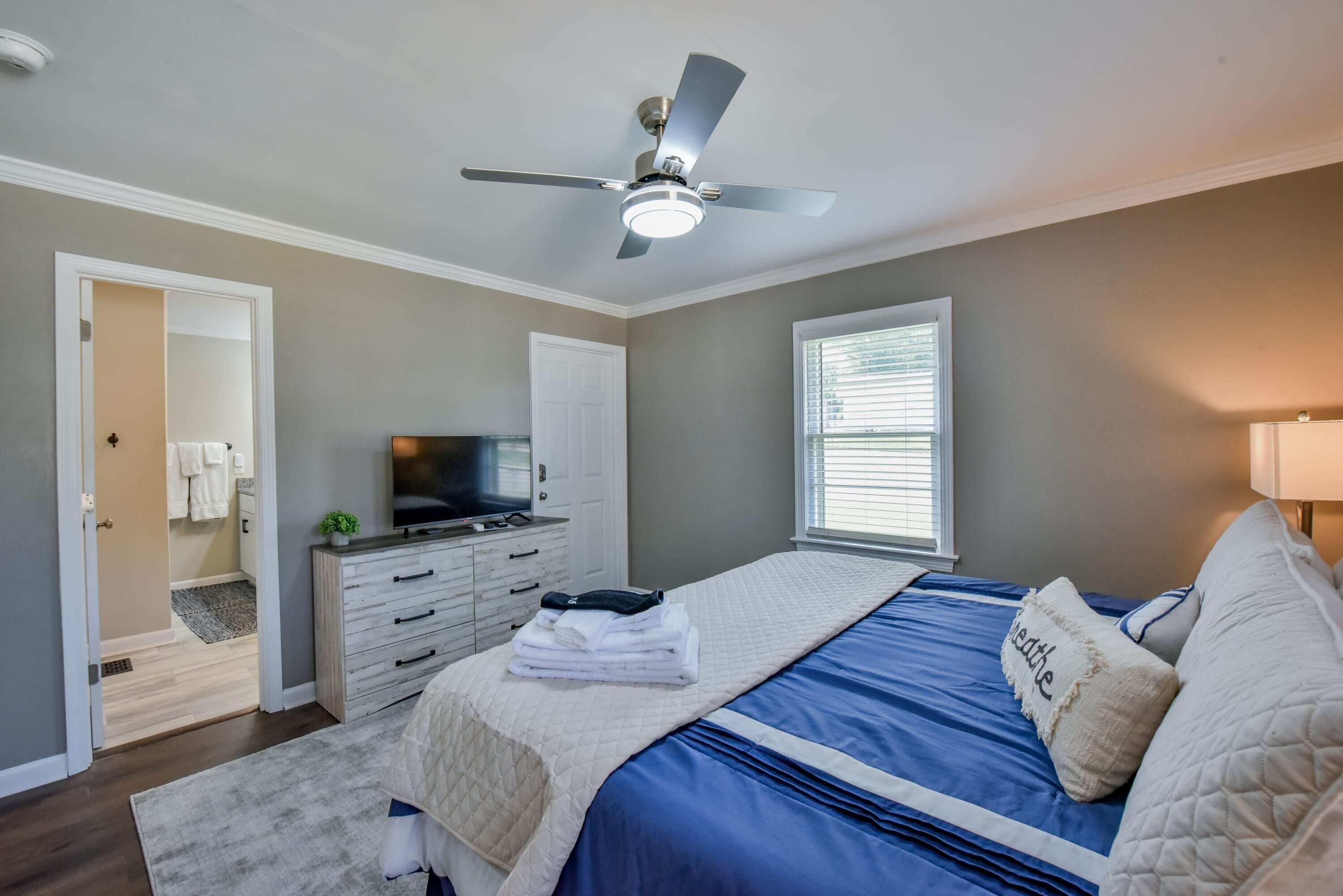 A bedroom with a bed, a ceiling fan, a TV, a dresser, and a window.