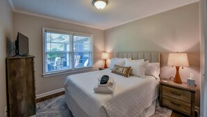 Un dormitorio ordenado con cama, lámparas de noche, mesita de noche, televisor en un mueble y una ventana con persianas.