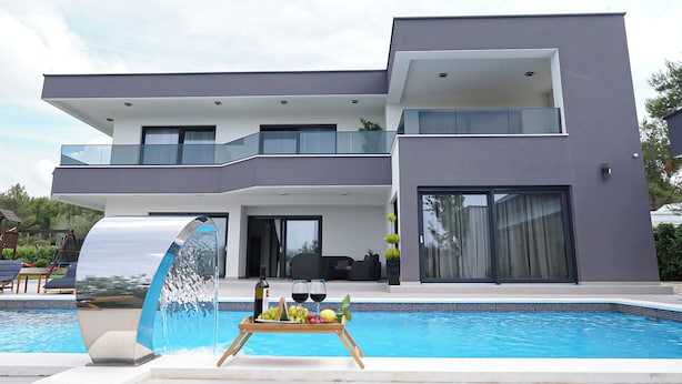A modern two-story house with a swimming pool, a dining area with outdoor seating, and a large glass window.