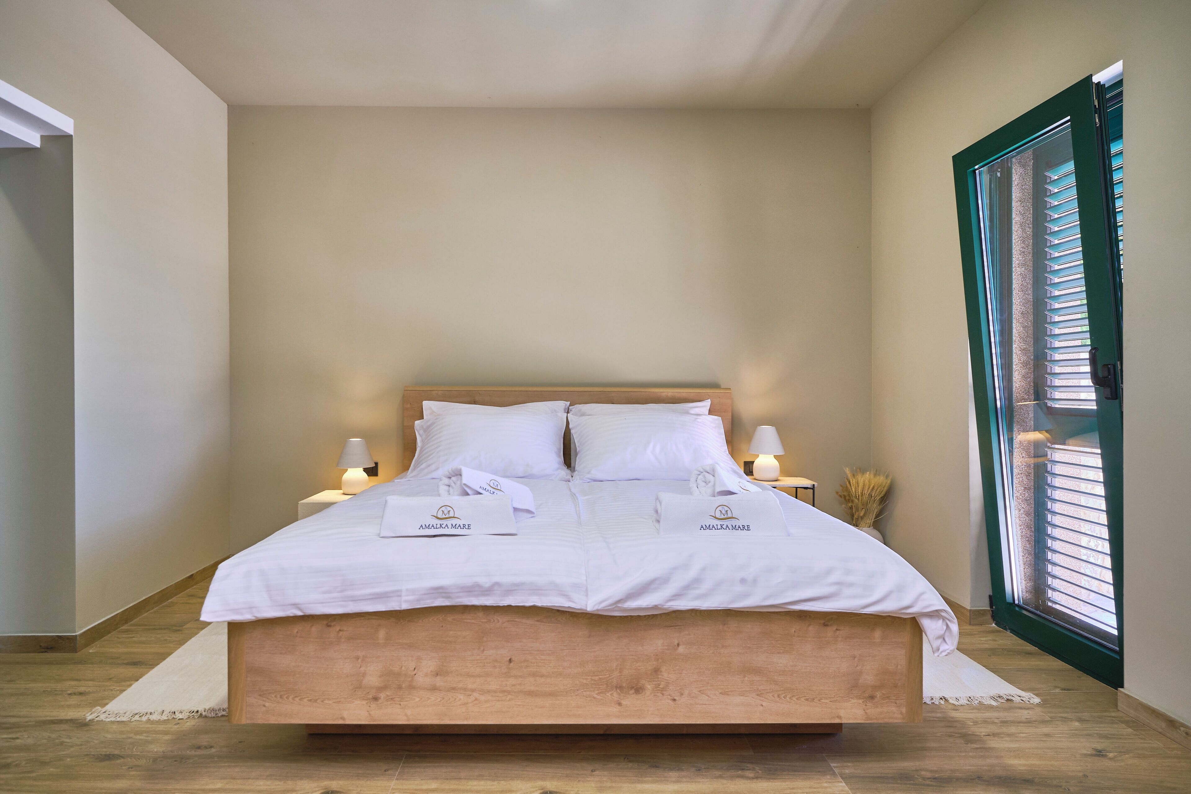 A neatly made bed with white linens and towels, a wooden headboard, bedside lamps, and a window with blinds.