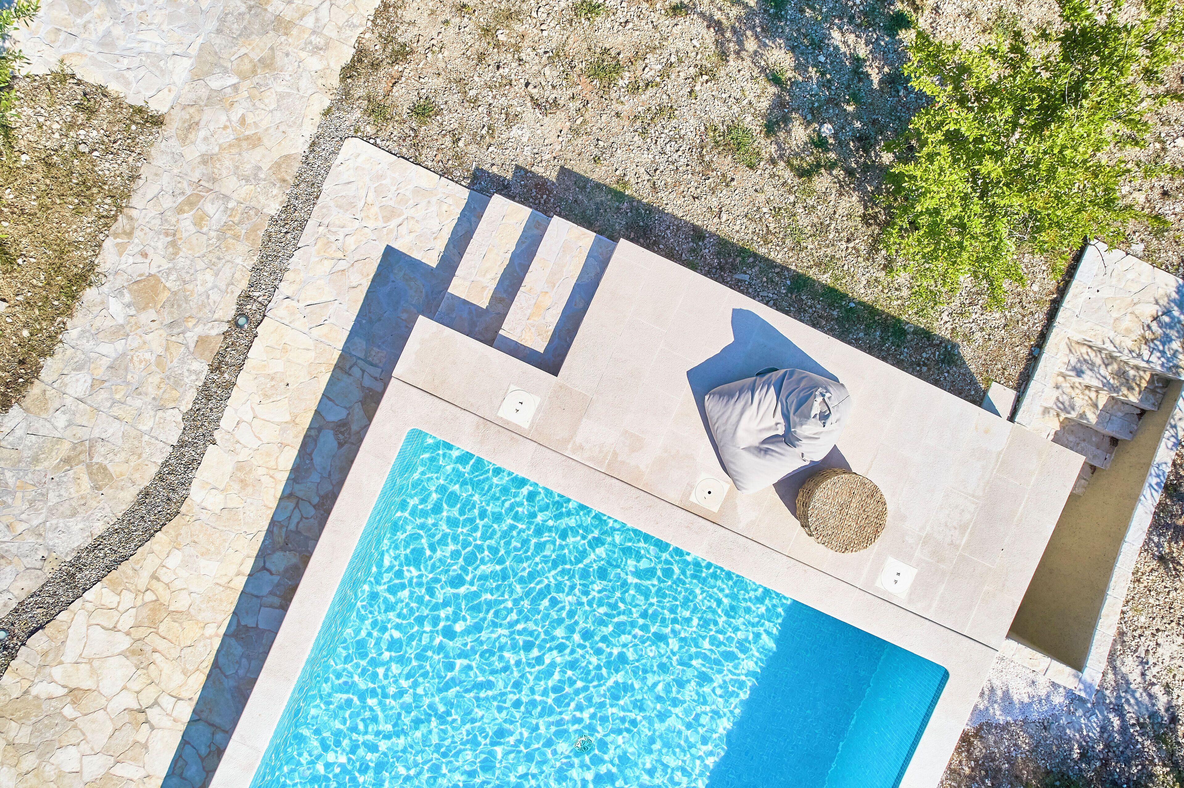 An aerial view of a swimming pool with a stone deck, a sun lounger, and a small garden area.