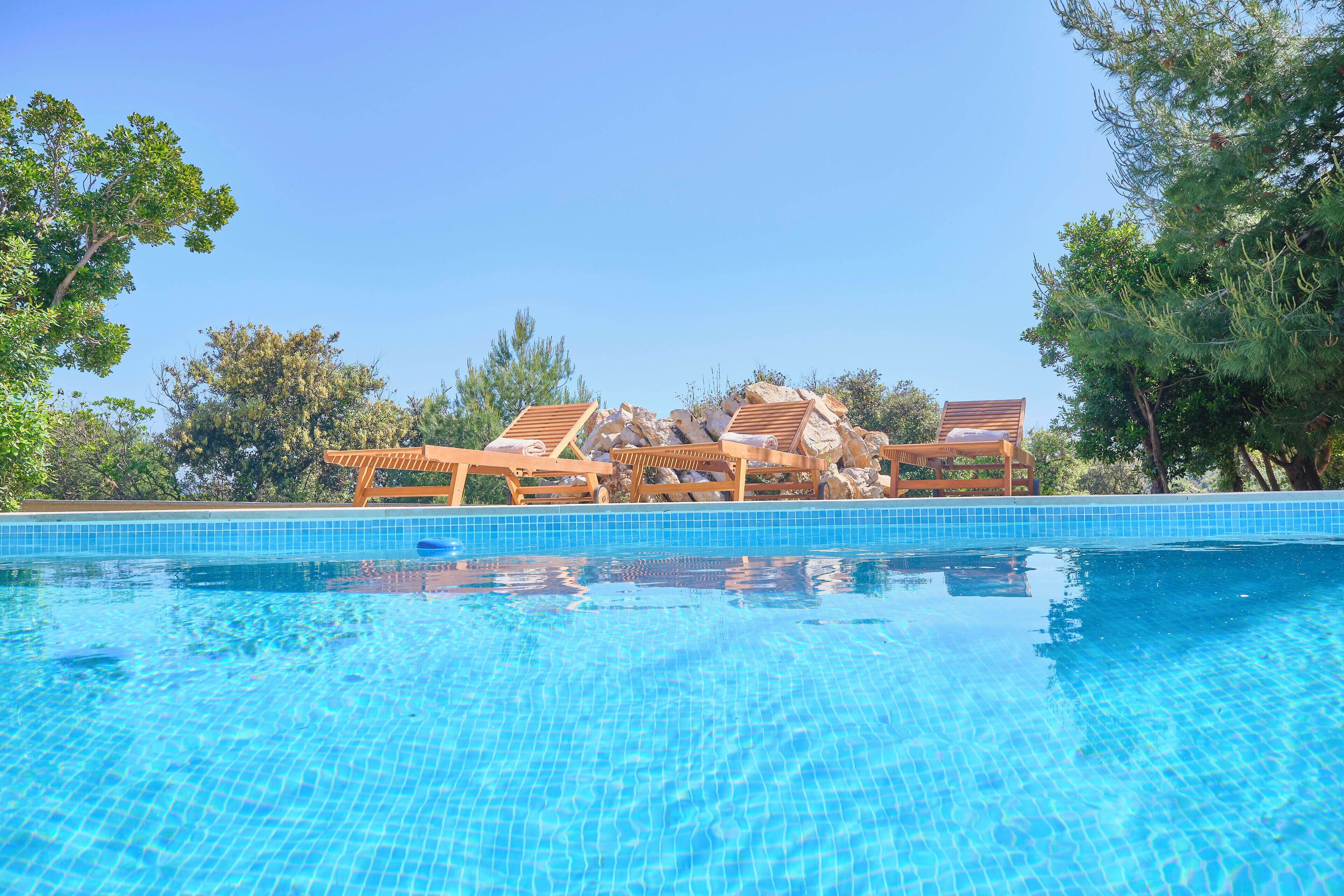 A clear blue swimming pool with wooden loungers and a stone structure in the background.