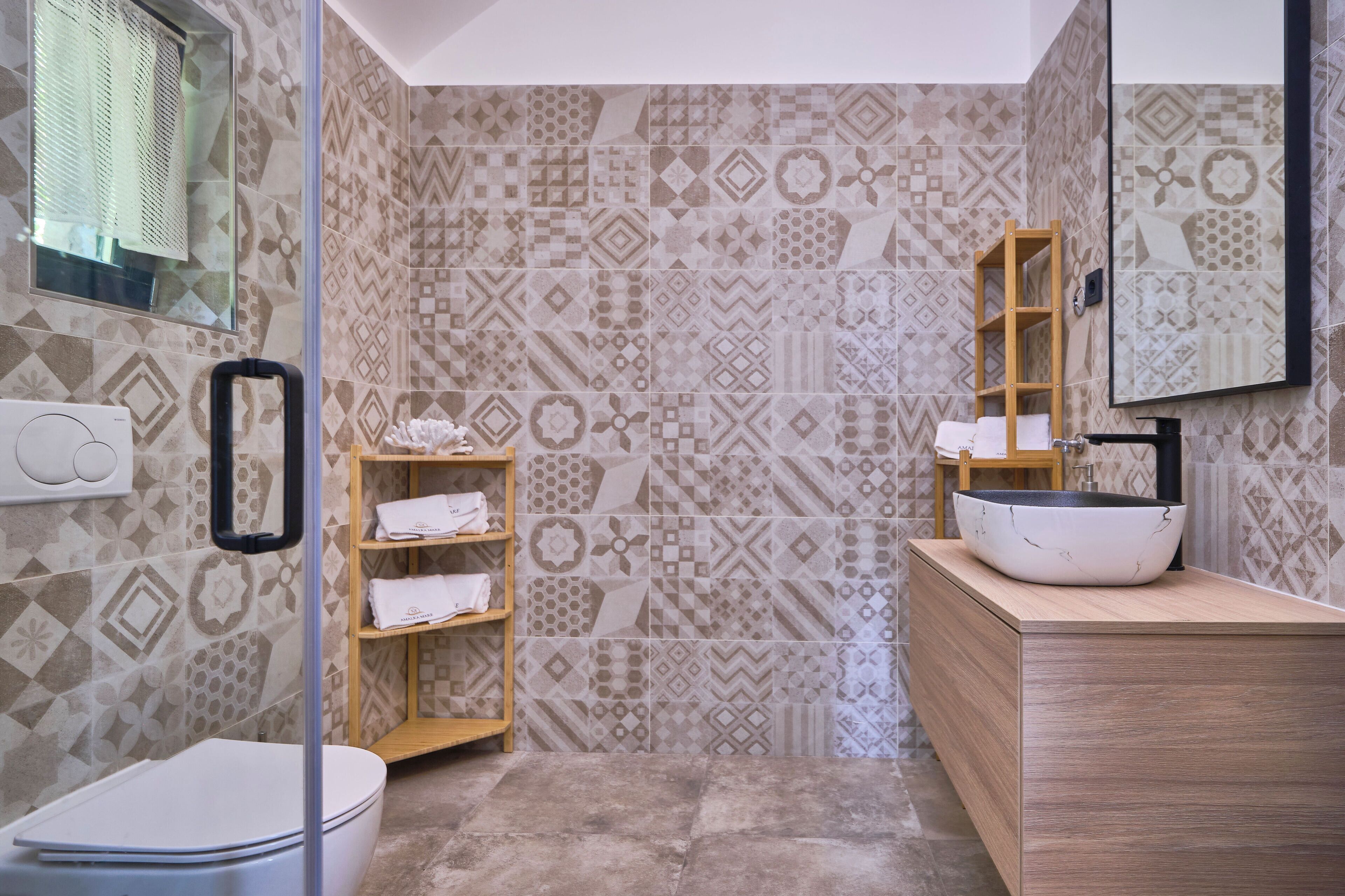 A bathroom with patterned tiles, a wooden shelf with towels, a white sink with a black faucet, and a mirror on the wall.