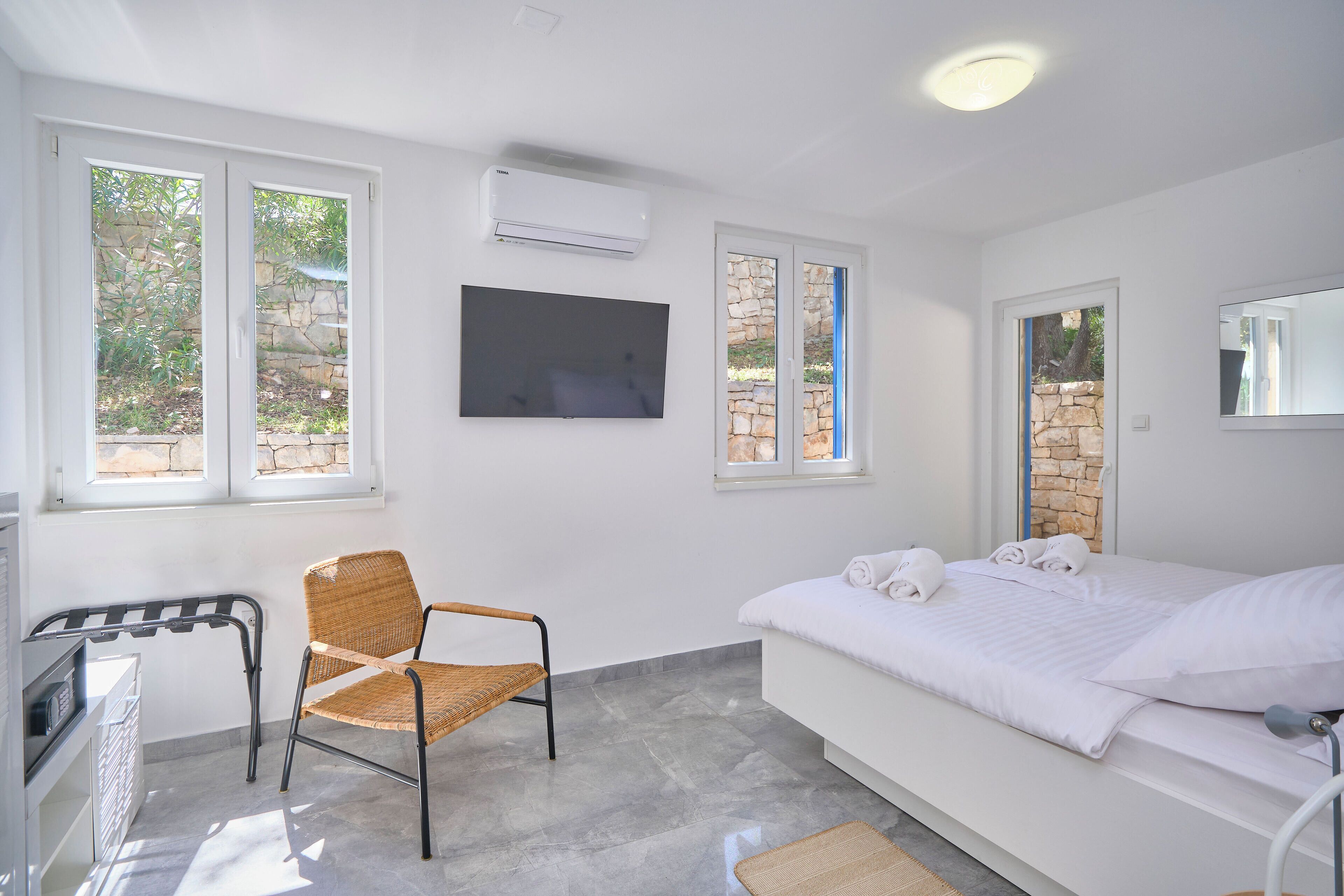 A bedroom with a bed, a chair, a TV, and a window with a stone wall view.