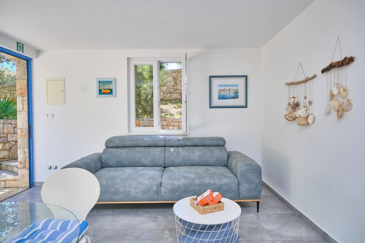 Ein modernes Wohnzimmer mit einem grauen Sofa, einem Glastisch und einem Korbtisch. An den Wänden hängen Bilder und durch das Fenster ist eine Steinwand zu sehen.