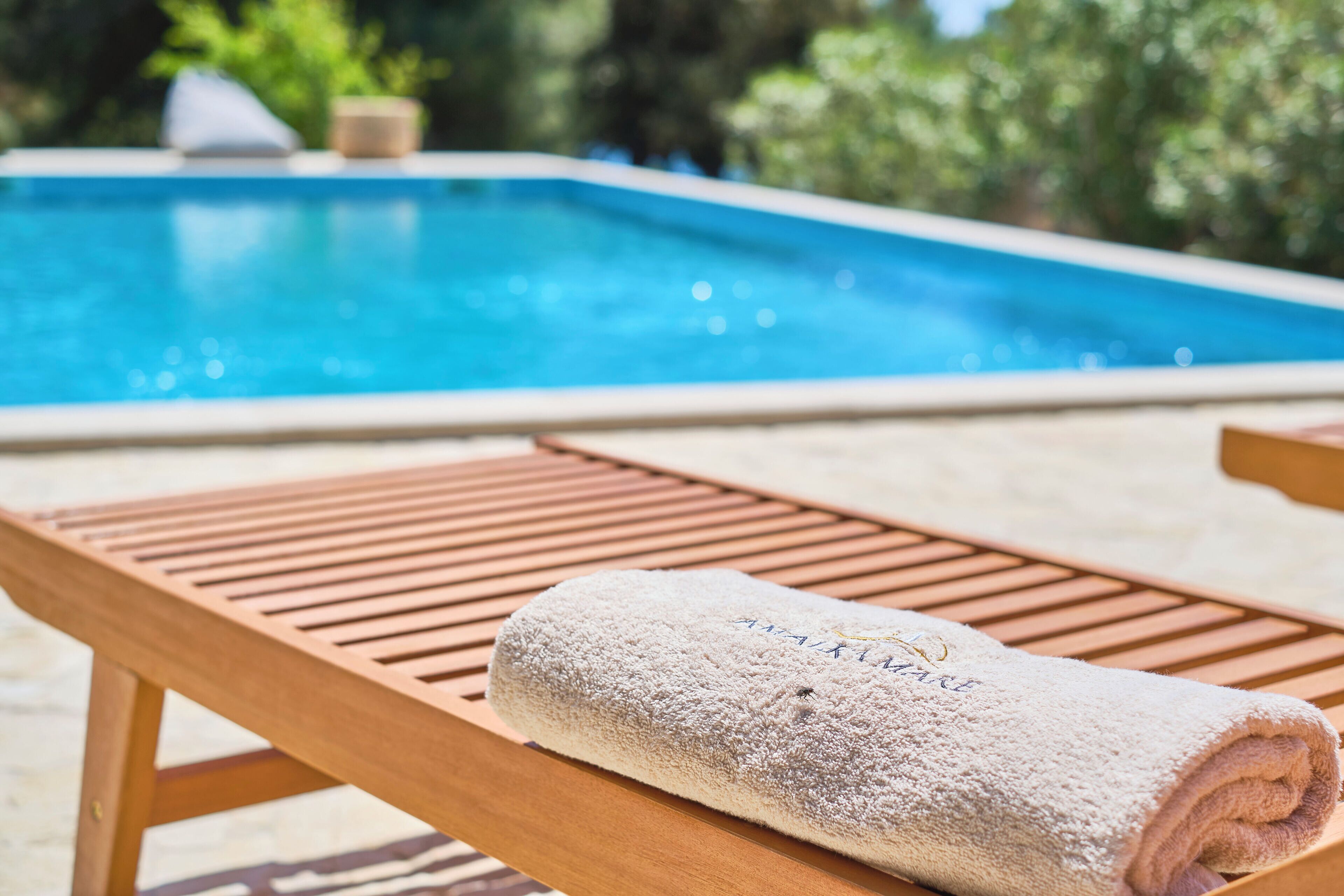 En solstol i trä med en hoprullad handduk på, en pool i bakgrunden och en naturlig utomhusmiljö.