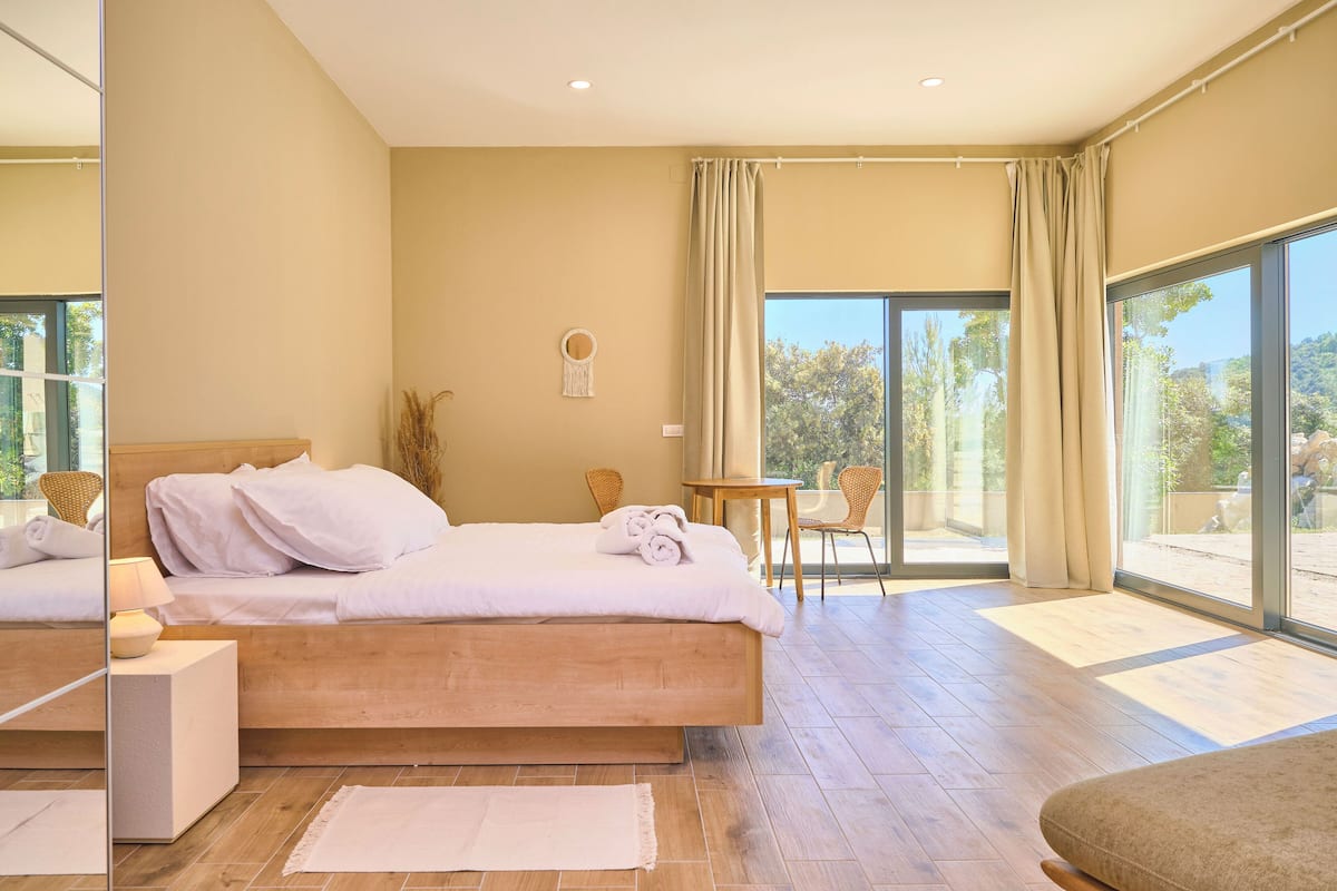 A modern bedroom with a large bed, a small table, and a chair near a sliding glass door leading to a balcony.