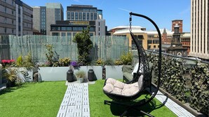 Un jardín en la azotea con una silla colgante, césped artificial, plantas en macetas y vistas a edificios de la ciudad.
