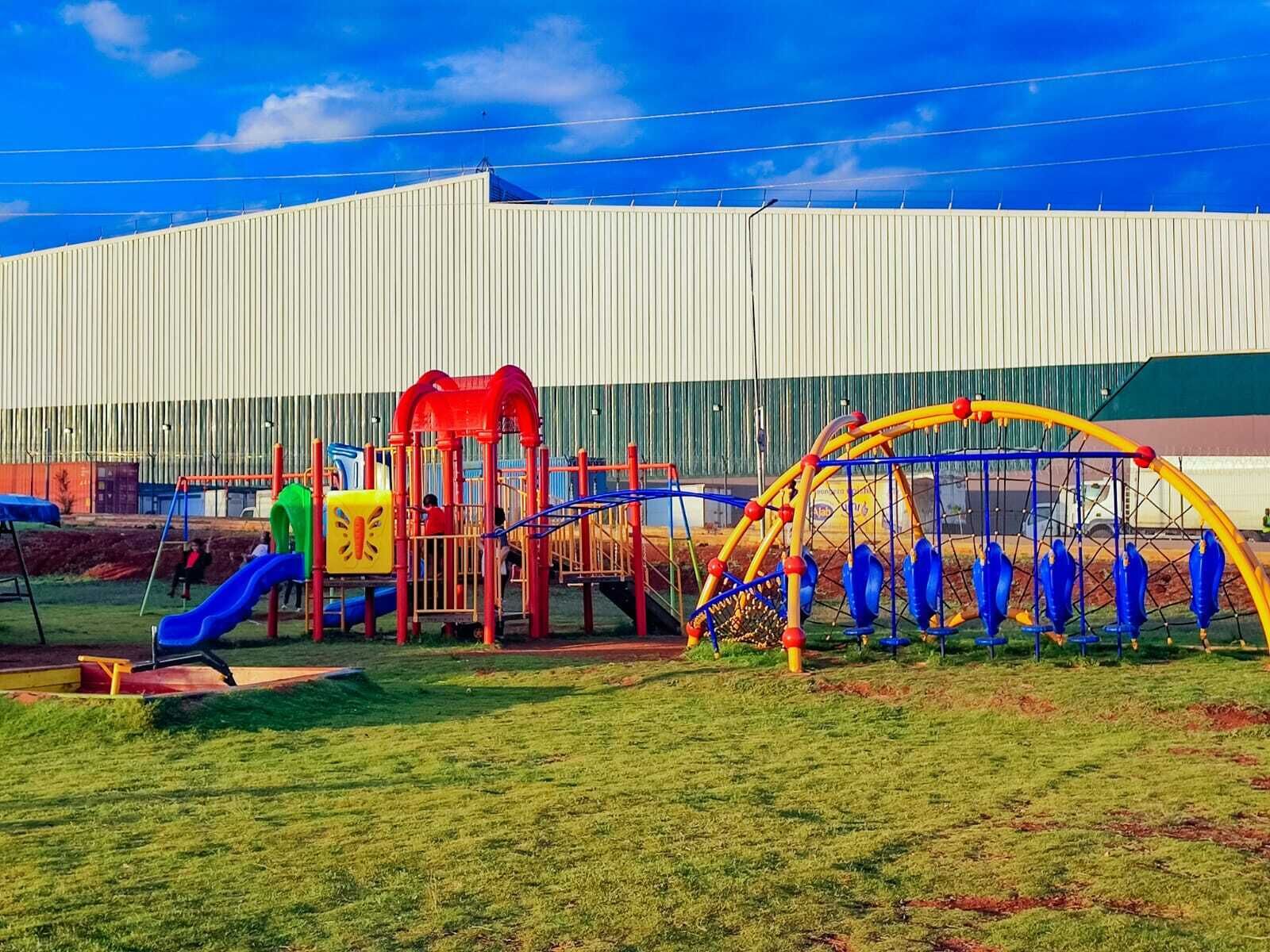 A playground with slides and climbing structures, set against a large building with vertical siding.