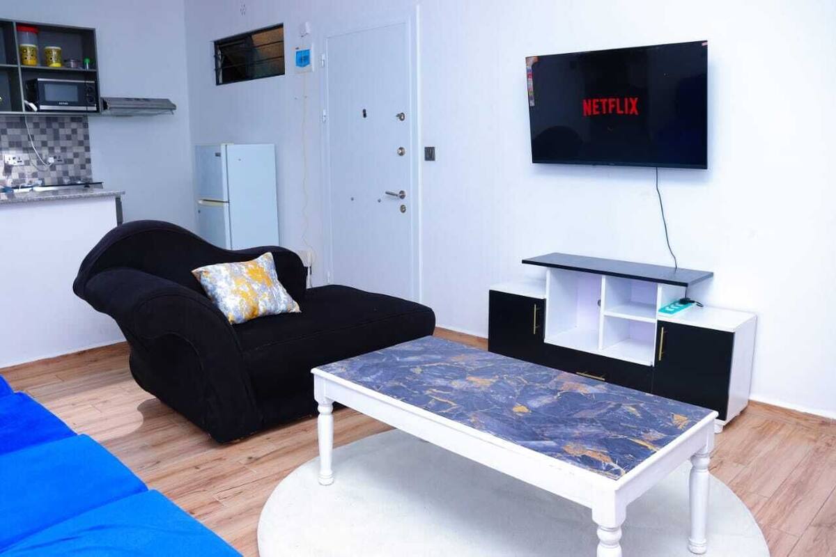 A compact living space with a black sofa, a white coffee table, a TV mounted on the wall, and a kitchenette area.