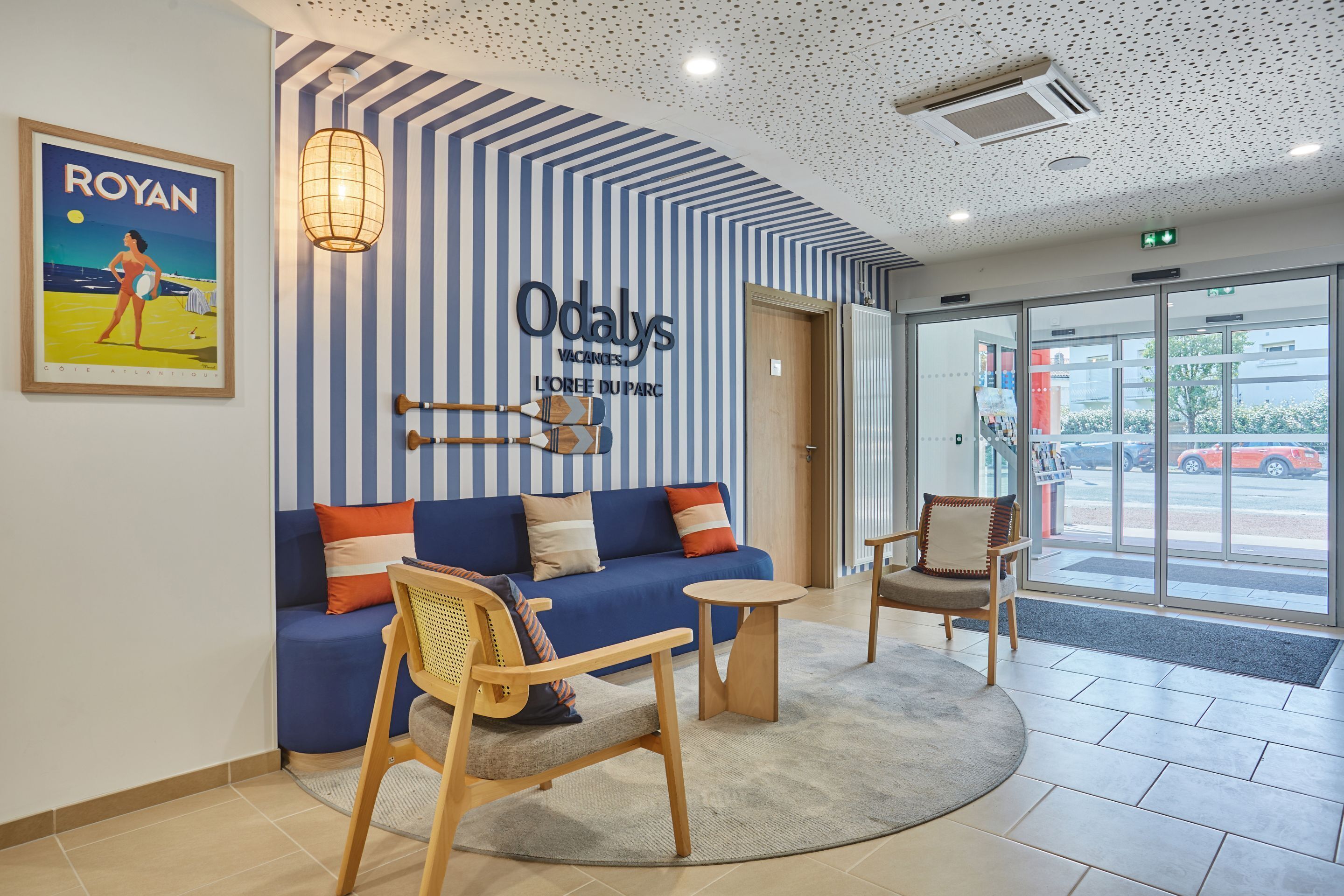 A reception area with a blue sofa, wooden chairs, a round table, and a wall sign reading "Odalys Vacances L'OEUVRE DU PARC".