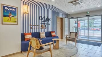 A reception area with a blue sofa, wooden chairs, a round table, and a wall sign reading "Odalys Vacances L'OEUVRE DU PARC".