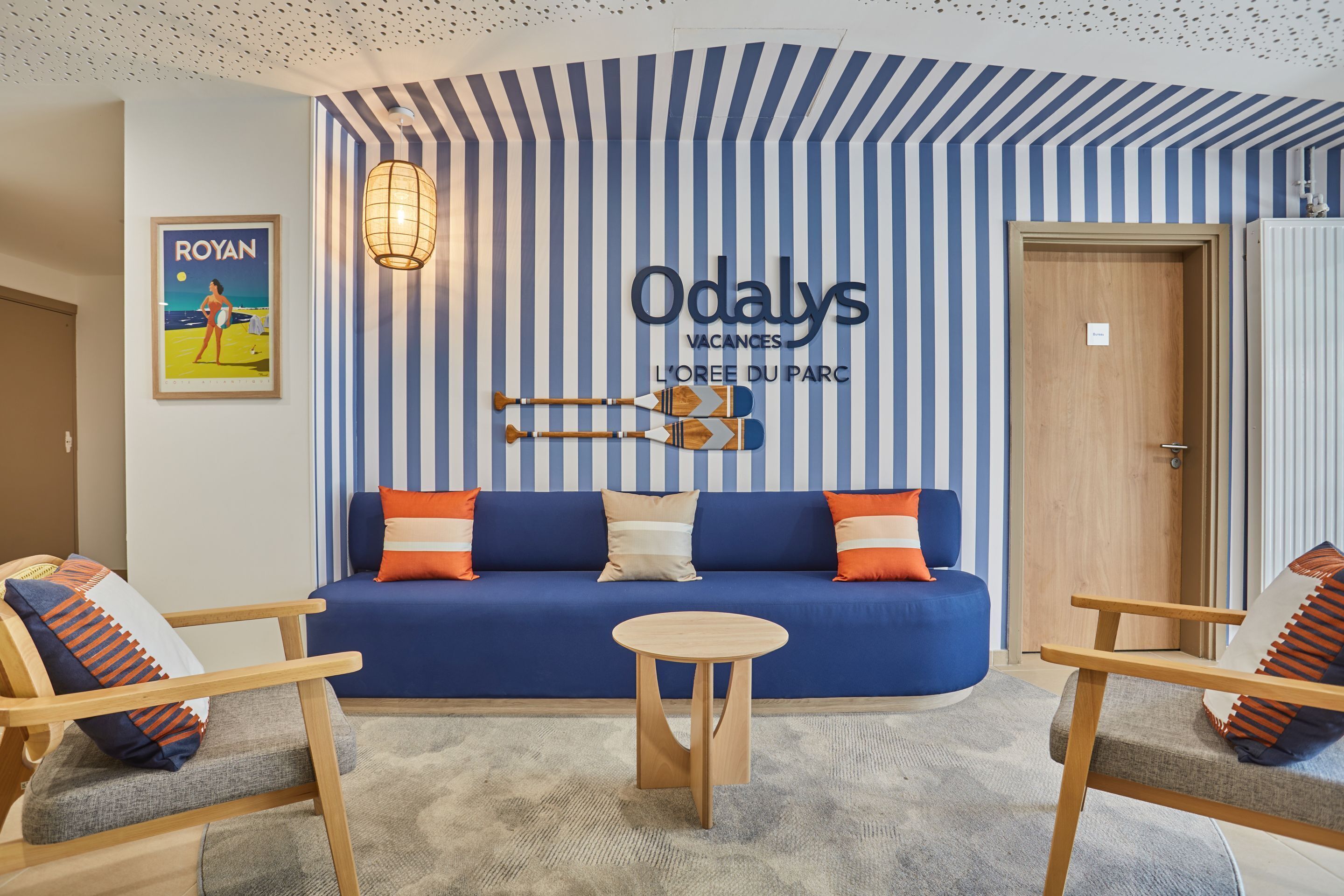 A room with a blue sofa, wooden chairs, a small round table, and a striped wall with a sign reading "Odalys Vacances L'Oeuf du Parc".