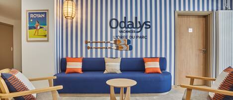A room with a blue sofa, wooden chairs, a small round table, and a striped wall with a sign reading "Odalys Vacances L'Oeuf du Parc".