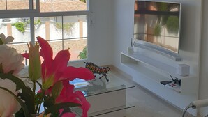 A modern living room with a large window, a flat-screen TV mounted on the wall, and a glass coffee table.