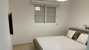 A bedroom with a bed, a window with blinds, a bedside table with a lamp, and a small potted plant.