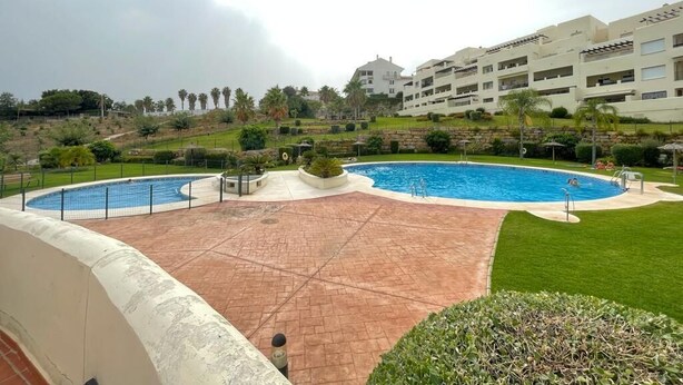 A swimming pool area with a paved deck, surrounded by greenery and a building with multiple balconies.