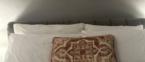 A neatly made bed with white bedding and a patterned pillow, framed artwork on the wall, and a grey wall.