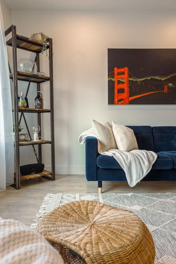 A living room with a blue sofa, a wooden shelf with decorative items, a painting of a bridge, and a woven basket.