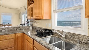 A kitchen with granite countertops, wooden cabinets, a stainless steel sink, and a view of the ocean through a large window. - OCS1 Ocean-Front w/ Panoramic, Water-Front Views (Nags Head)
