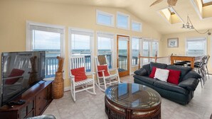 A spacious living area with a large flat-screen TV, a leather sofa, and a glass coffee table. There are multiple windows offering views of the ocean. - OCS1 Ocean-Front w/ Panoramic, Water-Front Views (Nags Head)