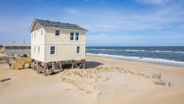 A multi-story beach house with a wooden deck, located on a sandy beach with ocean views. - OCS1 Ocean-Front w/ Panoramic, Water-Front Views (Nags Head)