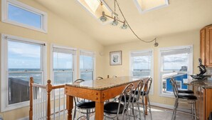 A dining area with a table and chairs, large windows with ocean views, and a ceiling light fixture. - OCS1 Ocean-Front w/ Panoramic, Water-Front Views (Nags Head)
