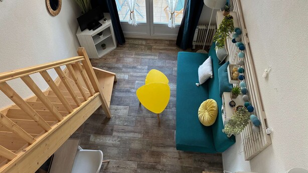 Een kamer met een houten trap, een blauwgroene bank met kussens, een gele stoel, een witte kast en uitzicht door een glazen deur.