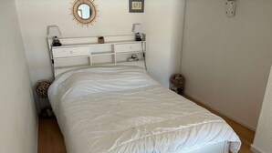 Een bed met witte lakens, een nachtkastje met een lamp, een spiegel aan de muur en een klein decoratief object.
