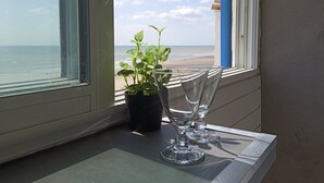 Une fenêtre donnant sur la plage, une plante en pot et deux verres vides posés sur un rebord.