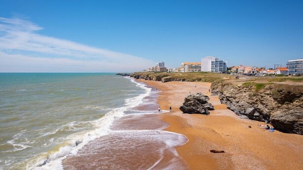 Una playa con costa rocosa, arena y edificios al fondo.