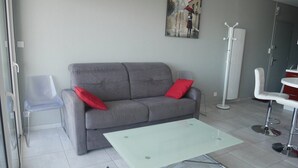 Een moderne woonkamer met een grijze bank, rode kussens, een glazen salontafel, een witte stoel en een kapstok.