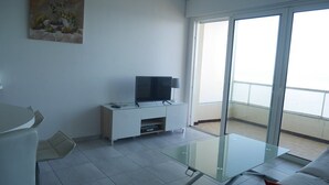 Een moderne woonkamer met een glazen salontafel, een flatscreen televisie op een witte kast, en een balkon met uitzicht.