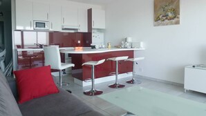 Een moderne keuken met witte kasten, een rood eiland en barstoelen. In de woonruimte staat een grijze bank met een rode kussen.