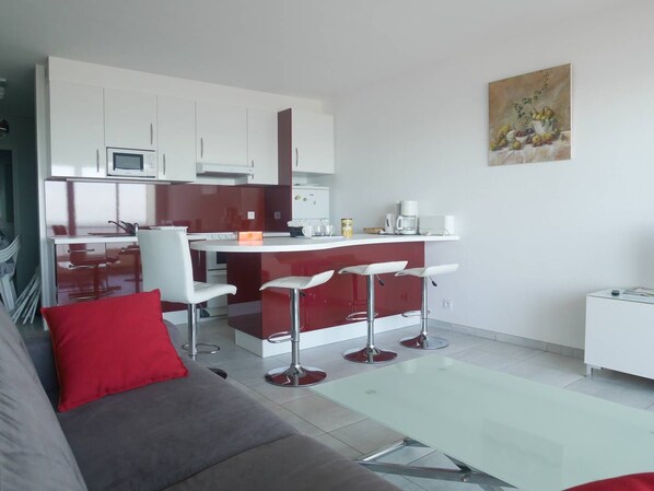 A modern kitchen with white cabinets, a red island, and bar stools. There is a gray sofa with a red pillow in the living area.