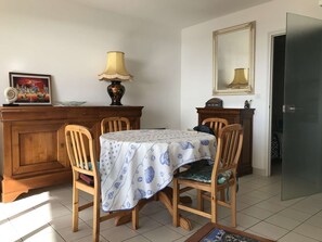 Uma sala de jantar com uma mesa coberta por toalha, cadeiras de madeira, um abajur, um quadro e um espelho na parede.