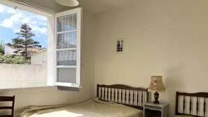 A small, single-bed room with a wooden headboard, a bedside table with a lamp, and a window with a view of a tree and buildings outside. - La tranquille - St Hilaire de Riez (SAINT HILAIRE DE RIEZ)