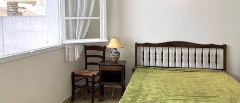 Una habitación con una cama, una silla, una mesita con lámpara, una ventana con cortinas e imagen colgada en la pared.