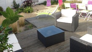 An outdoor patio with wicker furniture, a wooden deck, and a garden area with plants and a fence. - La Nantaise de la mer Saint Hilaire de Riez (SAINT HILAIRE DE RIEZ)
