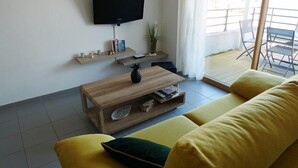 Ett modernt vardagsrum med en platt-TV som är monterad på väggen, ett träbord som används som soffbord och som är dekorerat med olika föremål, en gul soffa med gröna kuddar och utsikt över balkongen.