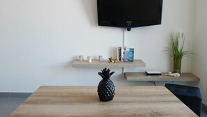 Ett vardagsrum med en platt-TV som hänger på väggen, ett träbord med en ananas som mittpunkt och en katt på en hylla.