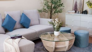 Una sala de estar moderna con un sofá gris claro, cojines azules, una mesa de centro de vidrio y un aparador blanco.