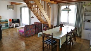 A spacious living area with a wooden staircase, a dining table with chairs, a sofa, and a television.