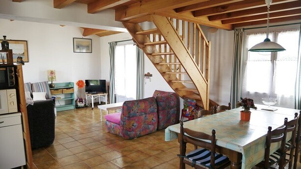 A spacious living room with a wooden staircase, a dining table with chairs, and a colorful sofa.
