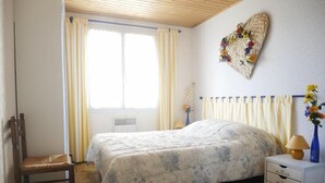 A bedroom with a bed, a chair, a bedside table with drawers, a radiator, and a window with curtains.