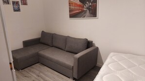 A small, modern room with a grey sectional sofa, a white mattress, and a framed picture of a red bus on the wall. - 2-bed Apartment Sleeps 7 With Wi-fi (London)
