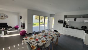 A modern kitchen with white cabinets, a dining table with a floral tablecloth, and a large sliding glass door leading to a patio. - La moderna de la pège - Saint hilaire de Riez (SAINT HILAIRE DE RIEZ)