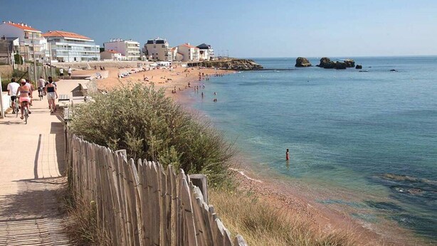 Ein Strand mit einer Holzbrüstung, klarem Wasser und Gebäuden im Hintergrund.