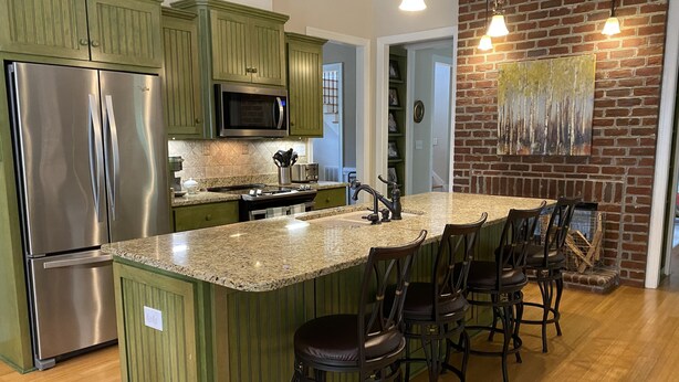 A kitchen with green cabinets, a stainless steel refrigerator, a built-in microwave, and a granite countertop. There are four bar stools and a hanging light fixture. - Masters Rental, 7 miles from course, 6 bedrooms, southern charm (Evans)